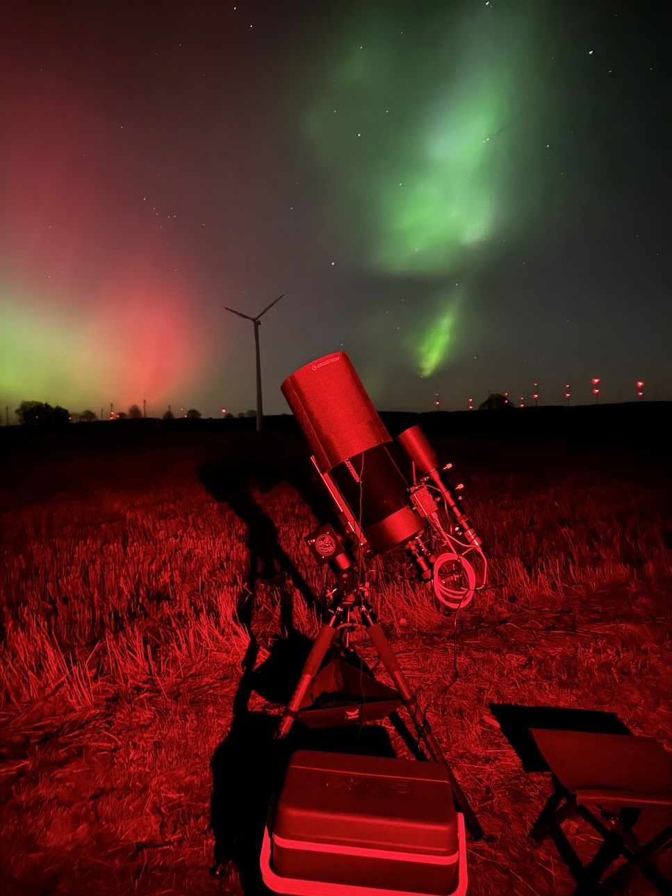 Teleskop im Rotlicht auf einem Feld, darüber grüne und rote Polarlichter